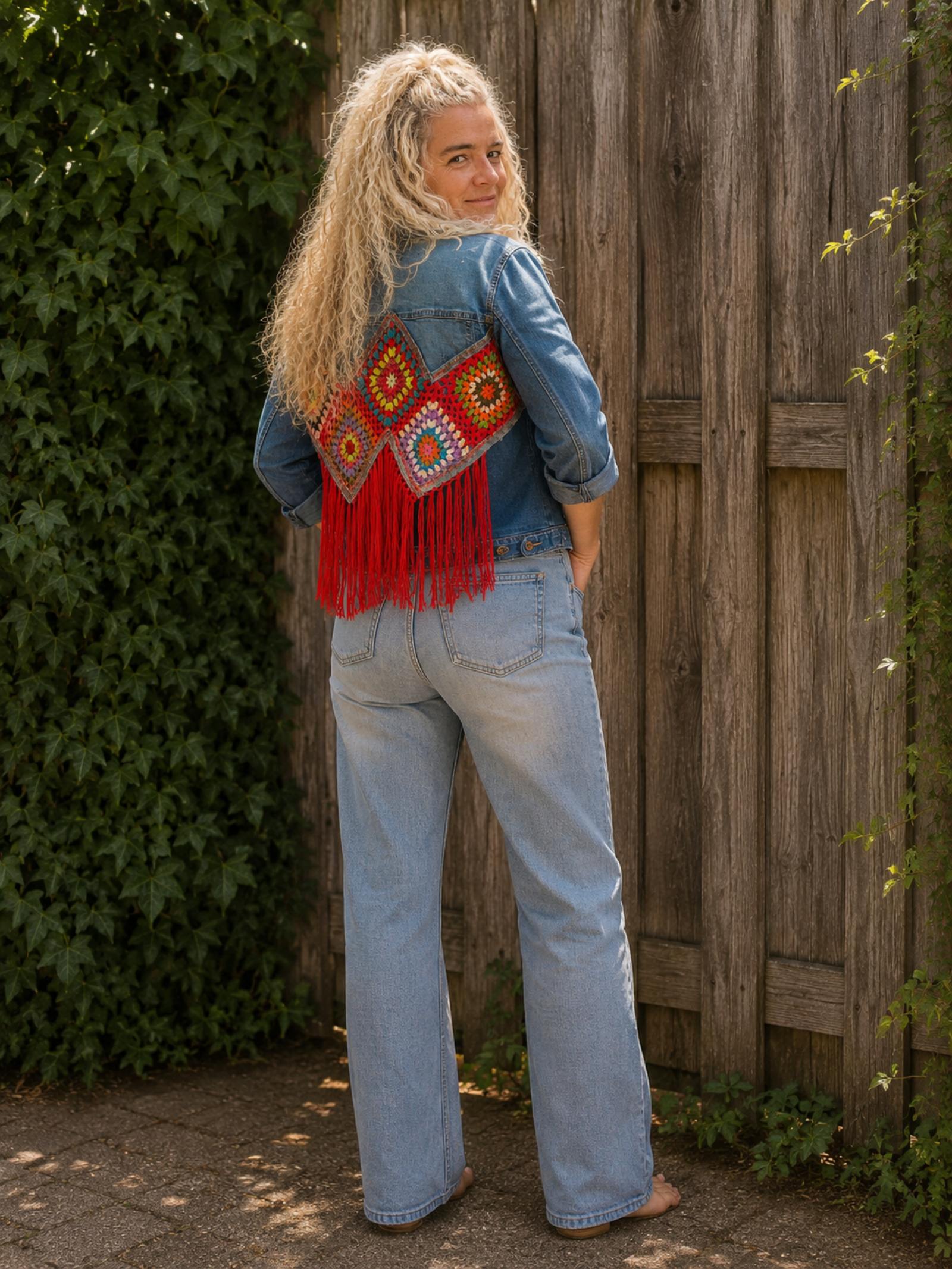 One of a Kind boho denim jasje van schuin achteren met kleurrijk gehaakt kunstwerk en rode franjes.
